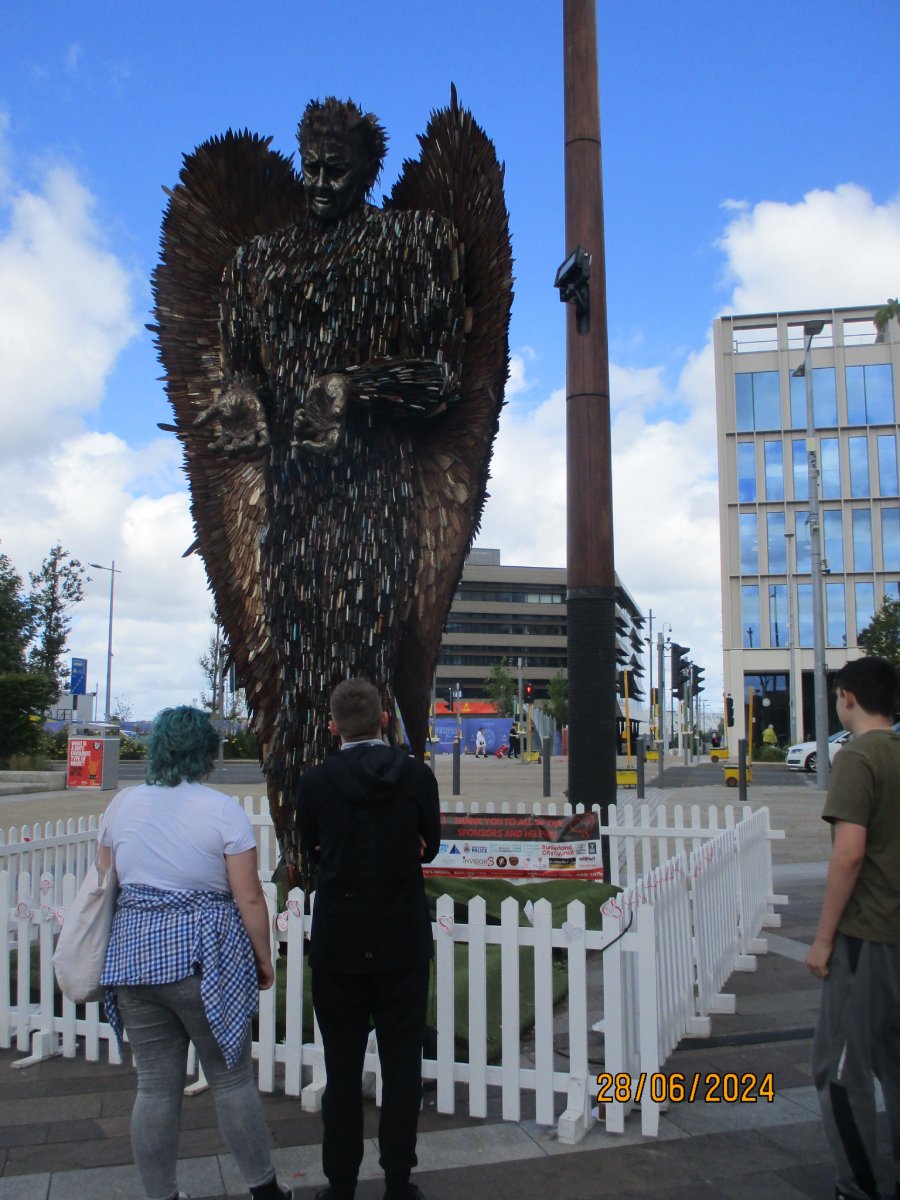 Three people viewing a large metal angel sculpture displayed in an outdoor public area.